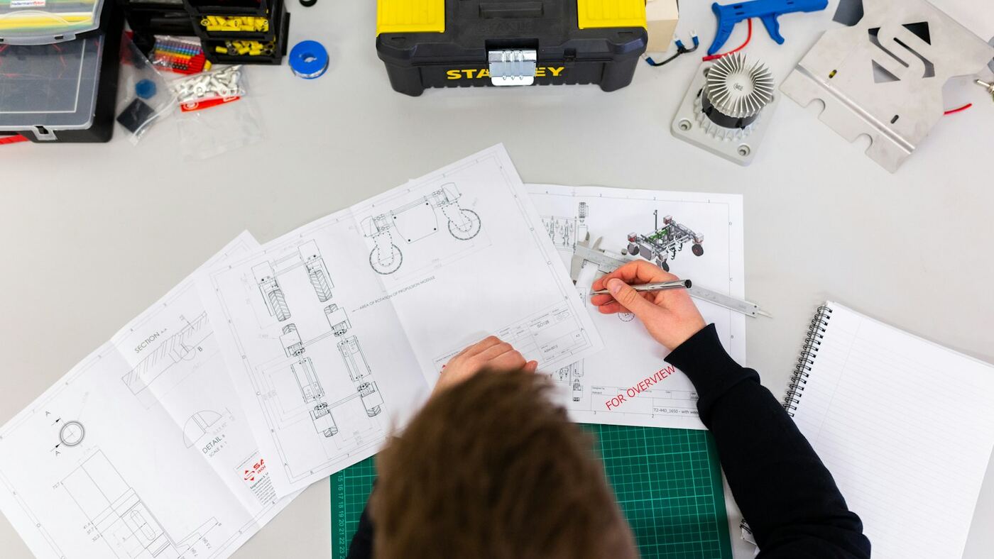 Overhead view of an engineer reviewing mechanical blueprints on a workbench with calipers, connectors, and a cutting mat